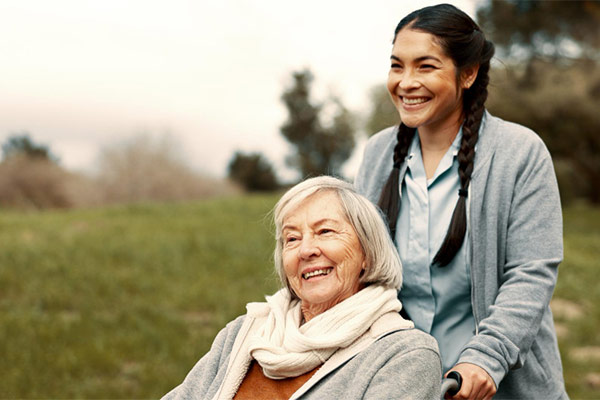 Two women, one sitting on a wheelchair, the other standing beside her, looking at each other with smiles.